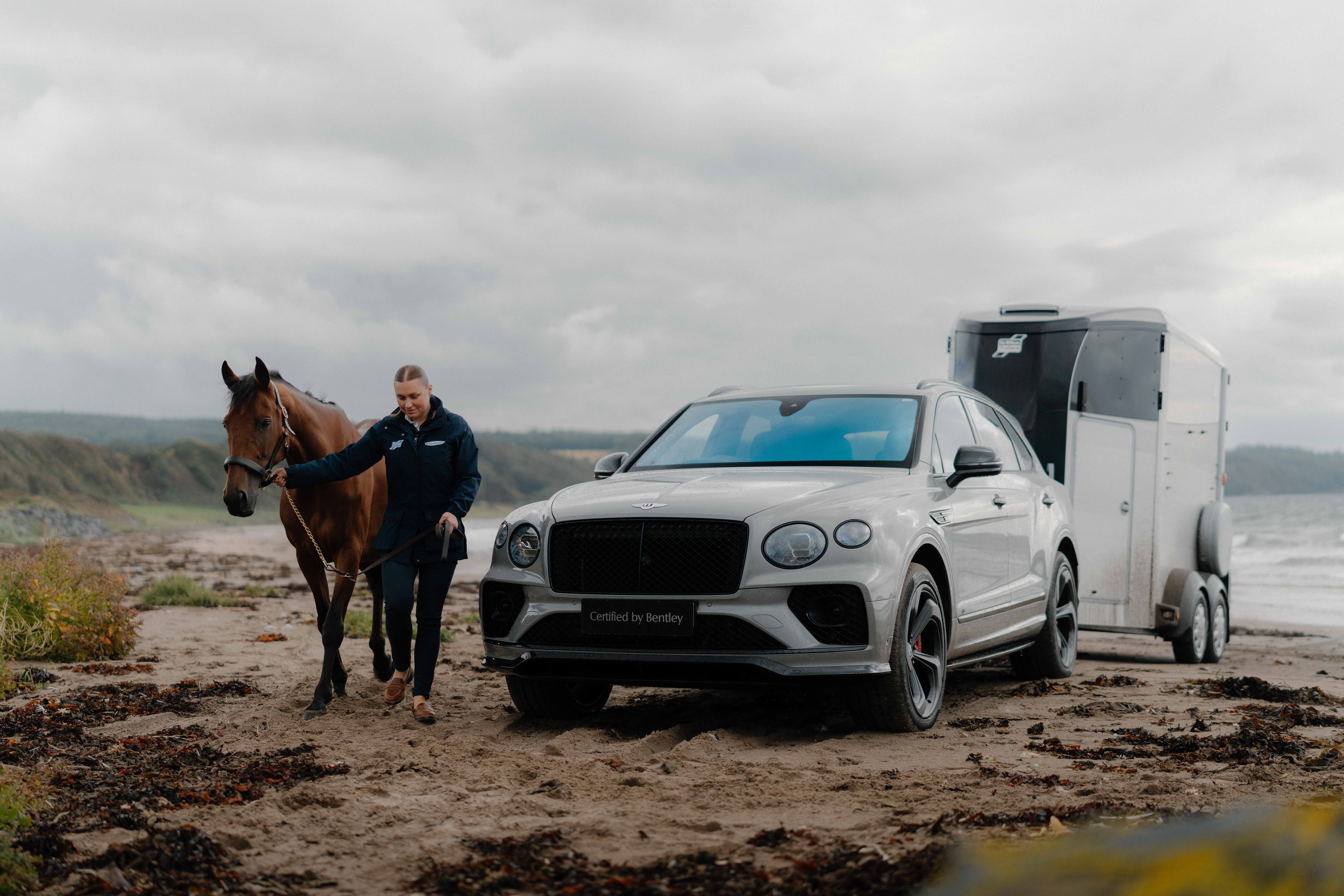 A woman with a horse stands next to a Bentley with a horse box attached to the back. They are on a beach